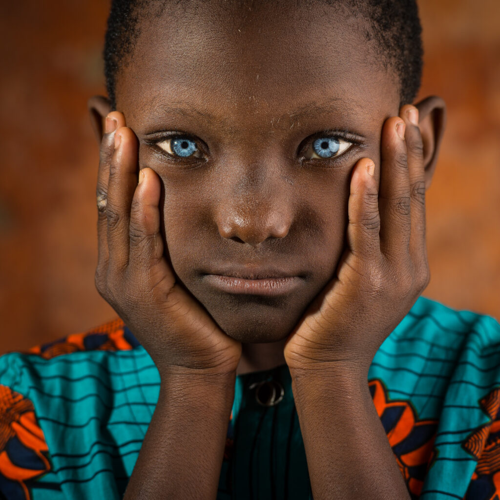 Un jeune garçon aux yeux bleus, le visage posé entre ses deux mains, en chemise de wax turquoise et orange, sur un fond ocre