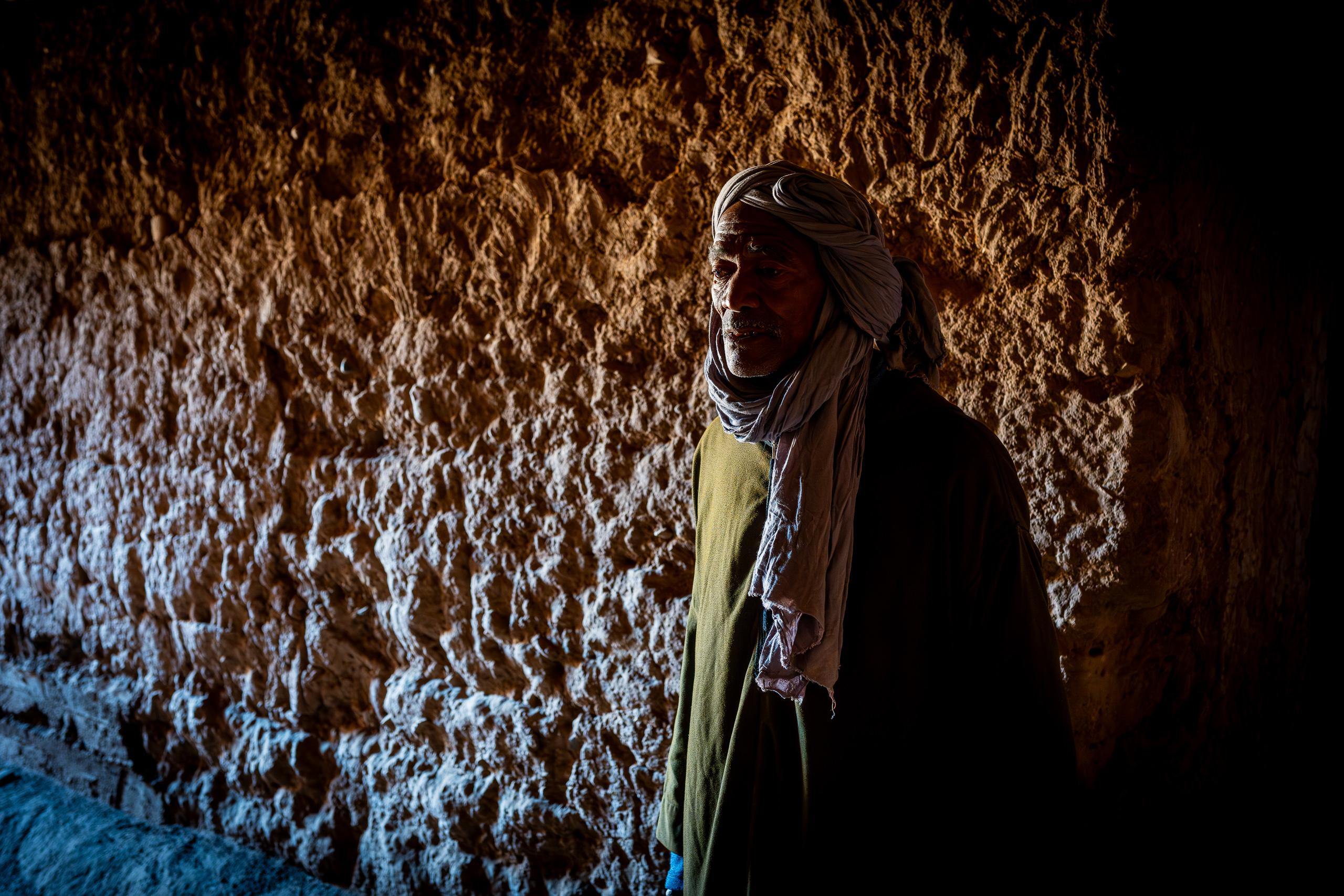 Un homme âgé à barbe grise, en djellaba kaki et chèche mauve, debout contre un mur de terre crue en lumière rasante